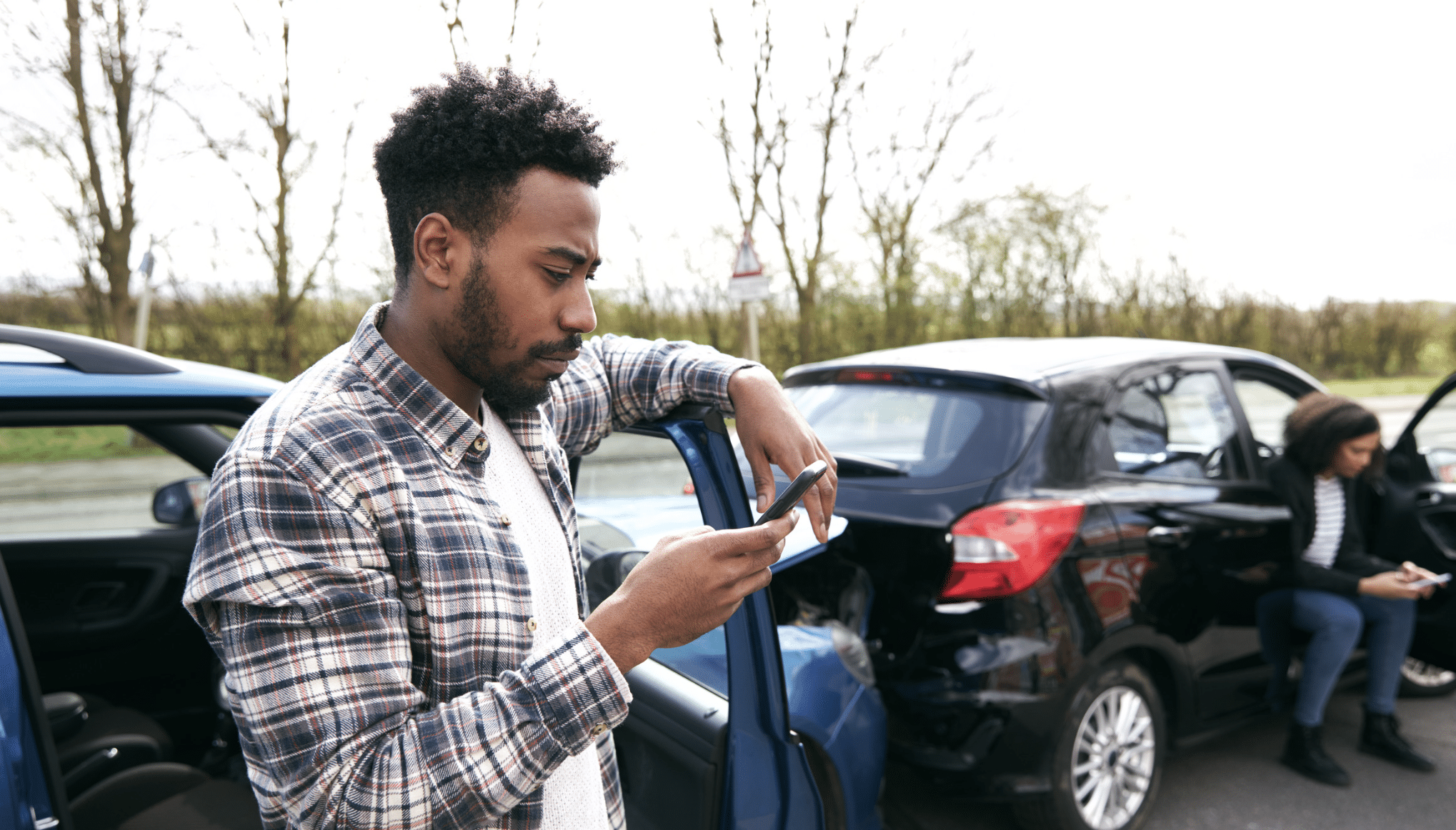 Man in a plaid shirt leaning on a blue car door and looking at his phone, with another person seated near another car in the background