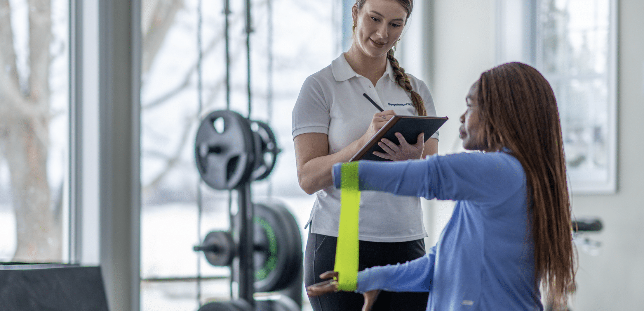 Physiotherapist takes notes on a clipboard while a client extends her arm with a neon-yellow strap in a gym setting.