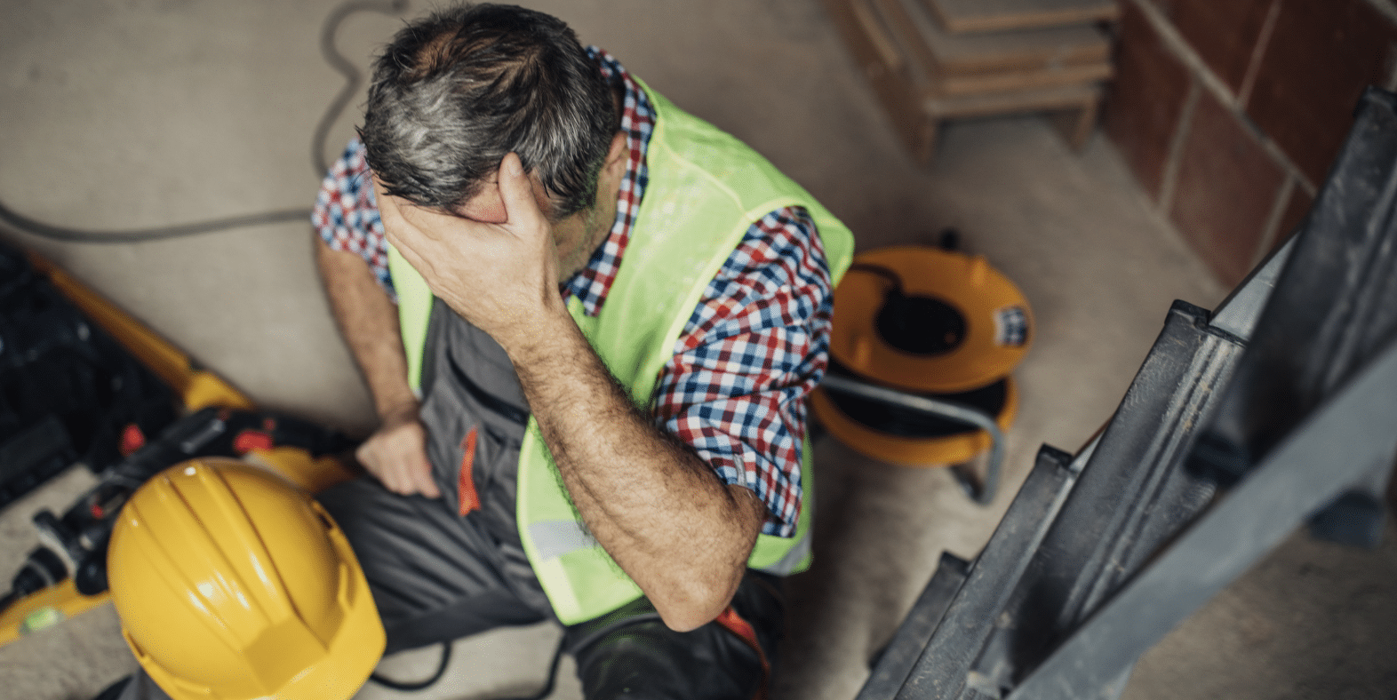 Construction worker in a neon safety vest sits on the floor, head in hand, with a yellow hard hat nearby at the worksite.