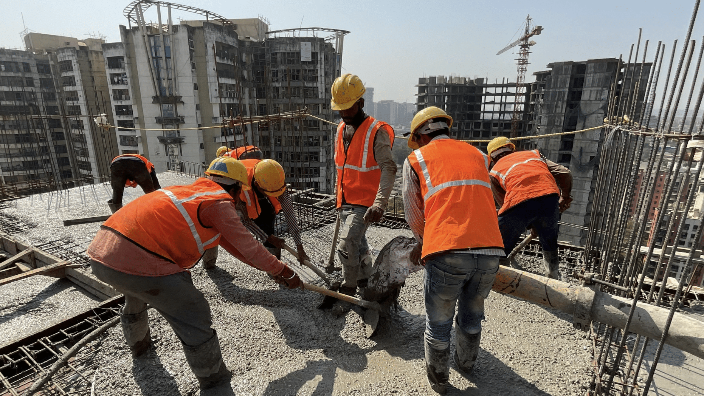 Construction workers in orange safety vests and hard hats spread wet concrete on a rooftop, with exposed rebar and a city skyline in the background.