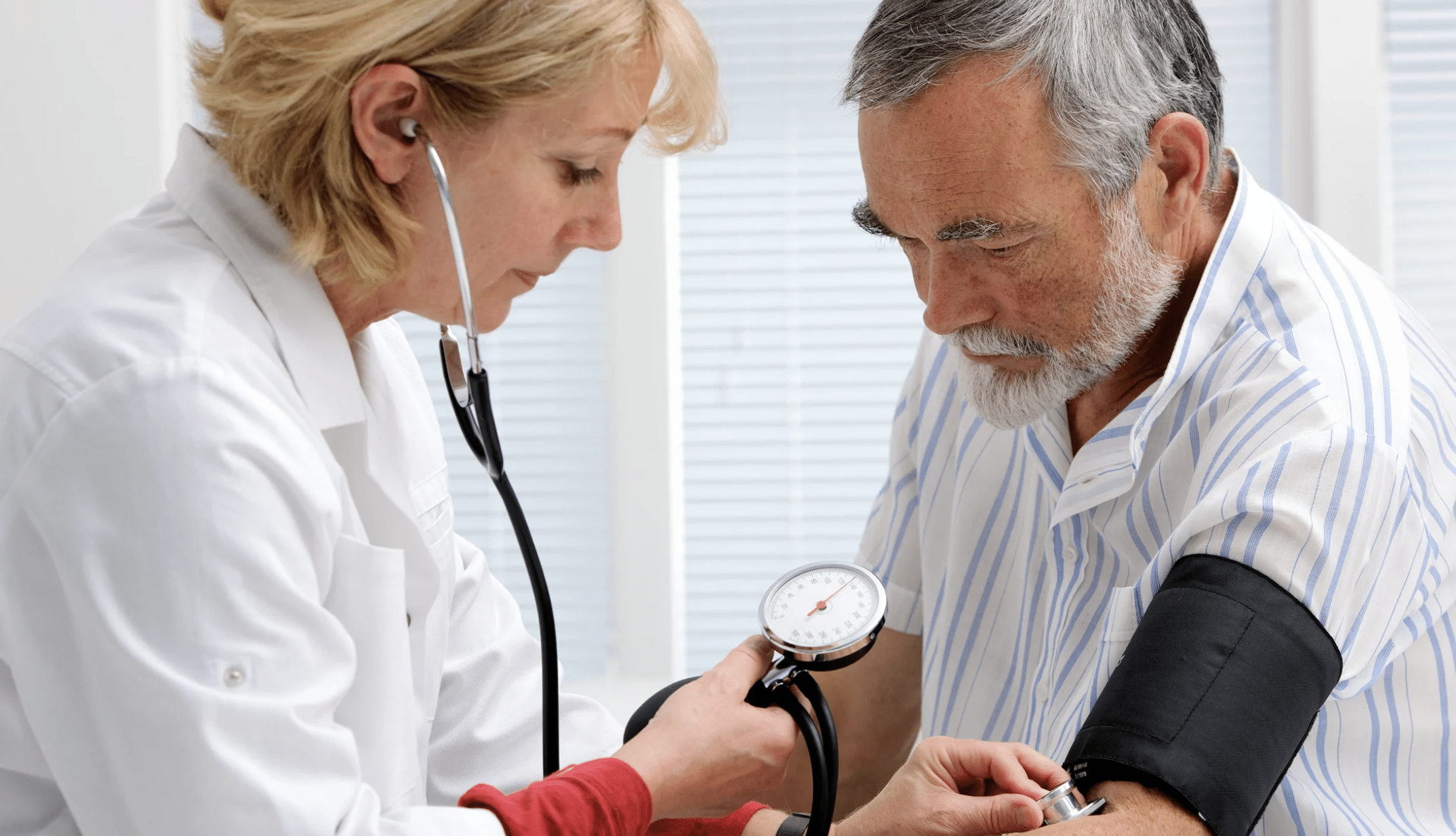 Healthcare professional checks an older man's blood pressure using a sphygmomanometer and stethoscope in a bright clinic.