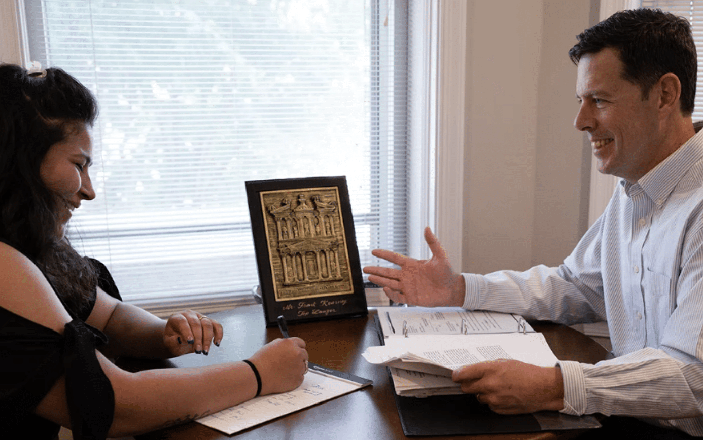 Two colleagues sit at a desk across from each other; one signs papers while the other gestures in conversation, with a framed picture on the table between them.