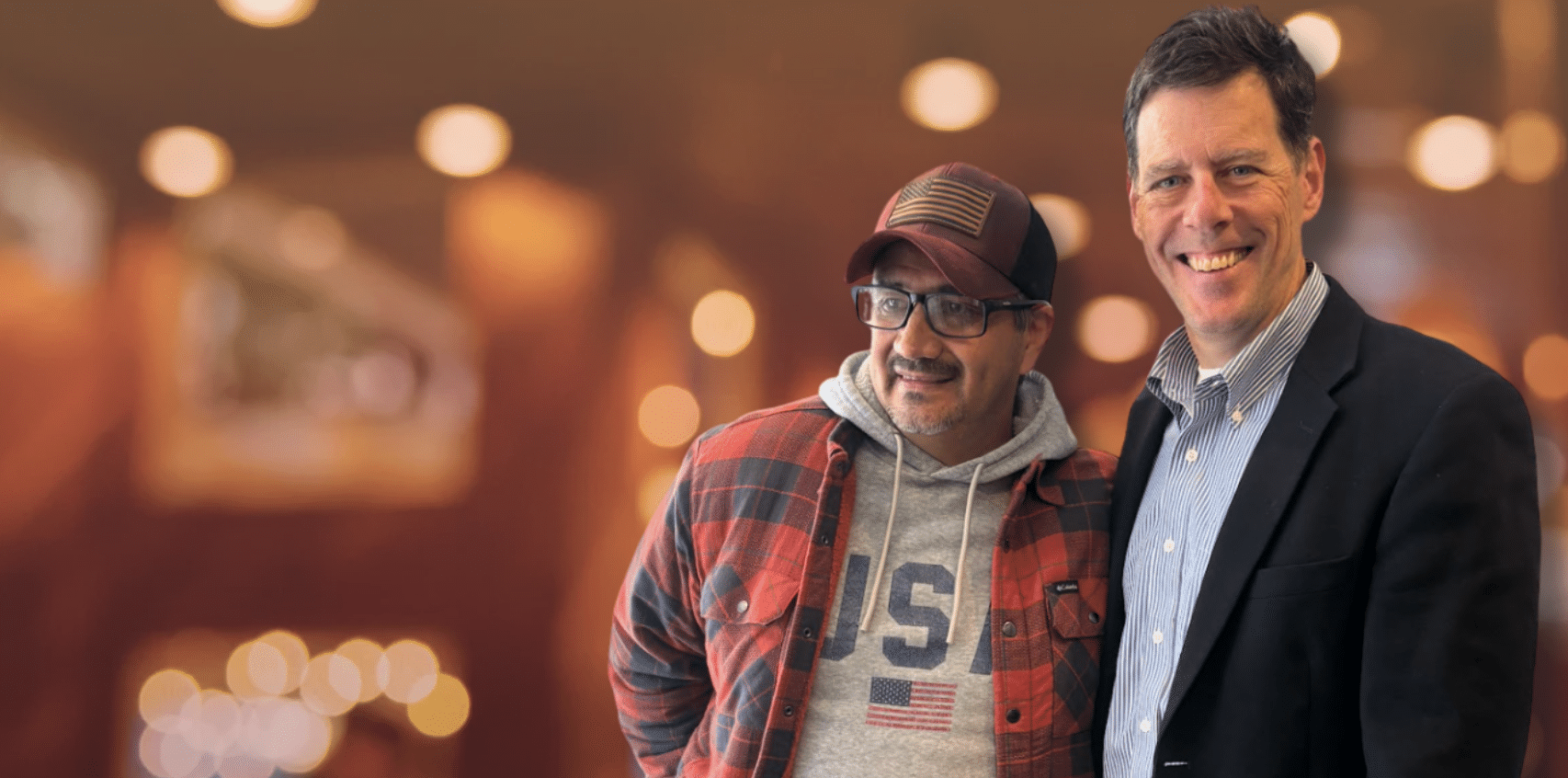 Two men smile at the camera in a warm, blurred indoor setting; one wears a red plaid shirt and cap, the other a blazer and striped shirt.