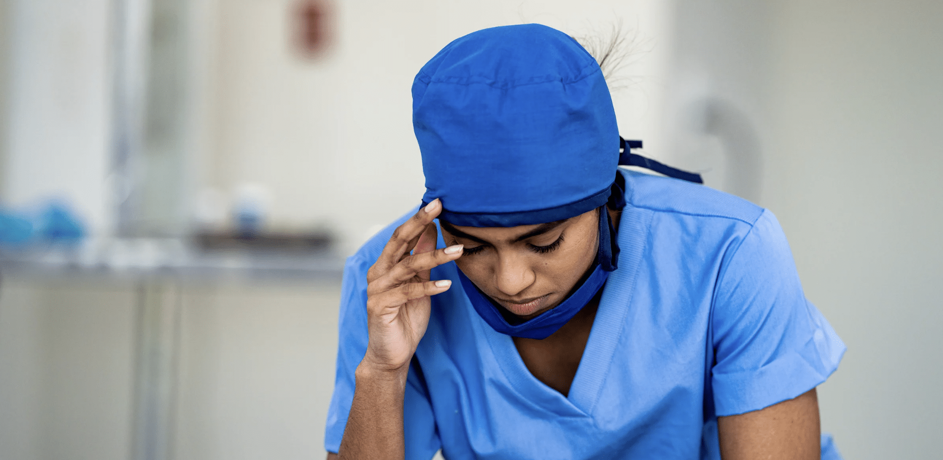 Female healthcare worker in blue scrubs with head covering, touching her temple and looking down in apparent fatigue or stress.